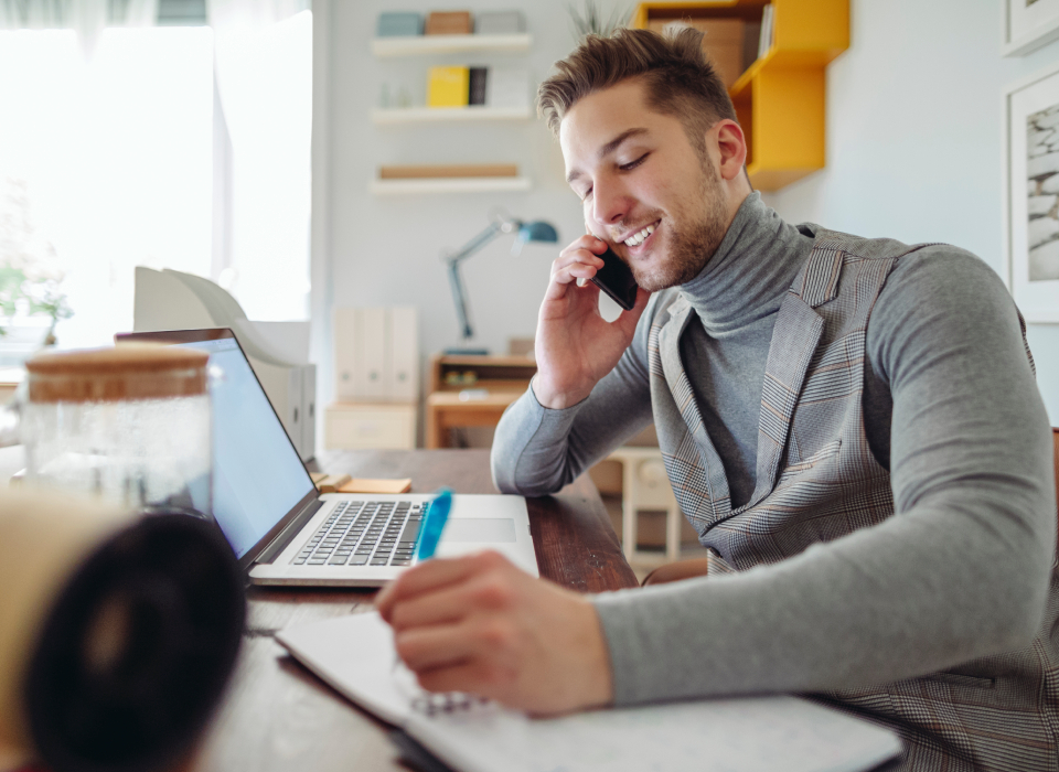 Junger Mann telefoniert am Schreibtisch mit Laptop und Notizbuch, konzentriert und freundlich.
